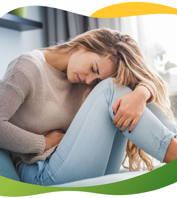 A young woman suffering from period bloating and sitting cramped on the floor holding her abdomen with her left hand