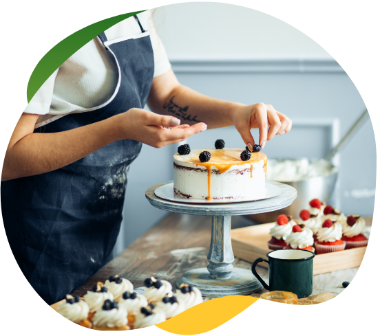 A person, standing in the kitchen, is decorating a cake with berries. On the work surface are several cupcakes